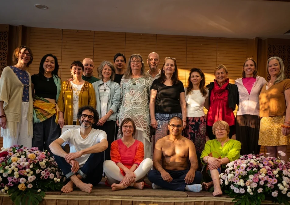 Group photo of yoga retreat participants and teachers smiling together indoors surrounded by flower arrangements